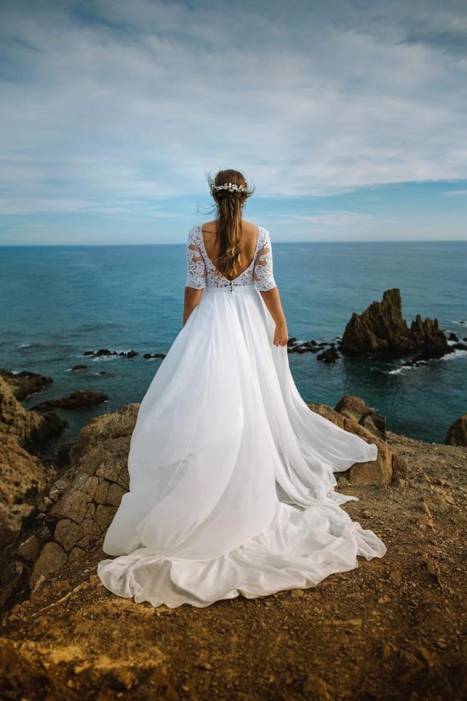 Bride standing on the rocks by the sea, her gown blowing in the wind