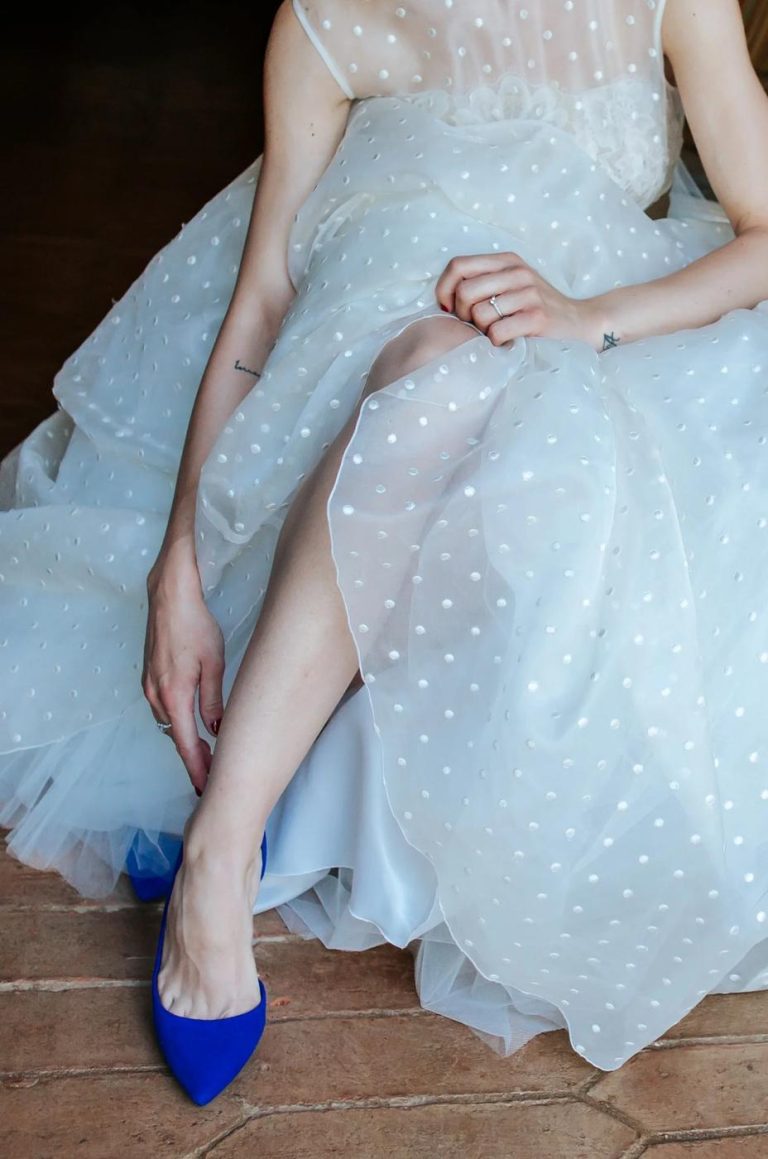 Detail of a bride slipping on blue wedding shoes beneath a polka-dot tulle gown